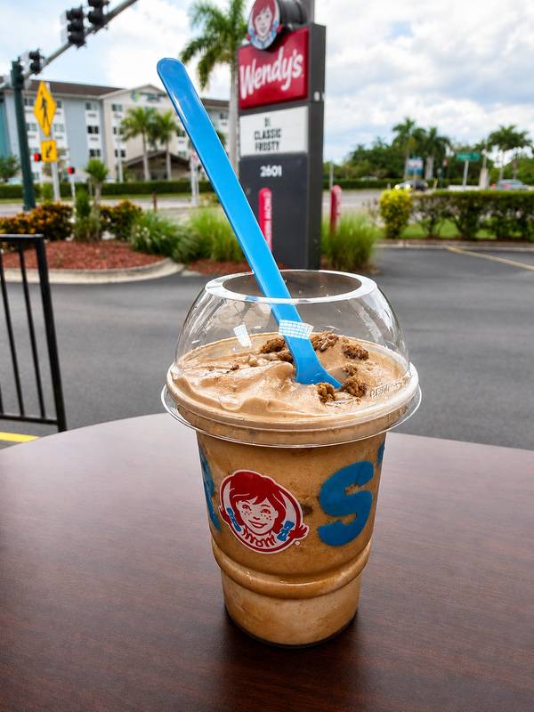 A Wendy's Frosty cup with a blue spoon, photographed in front of a Wendy's restaurant sign